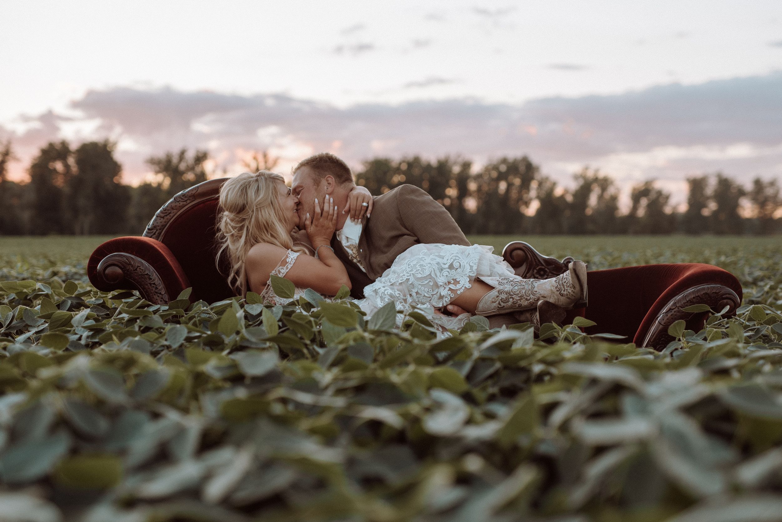 Newlyweds kissing on a vintage velvet sofa in a field at sunset